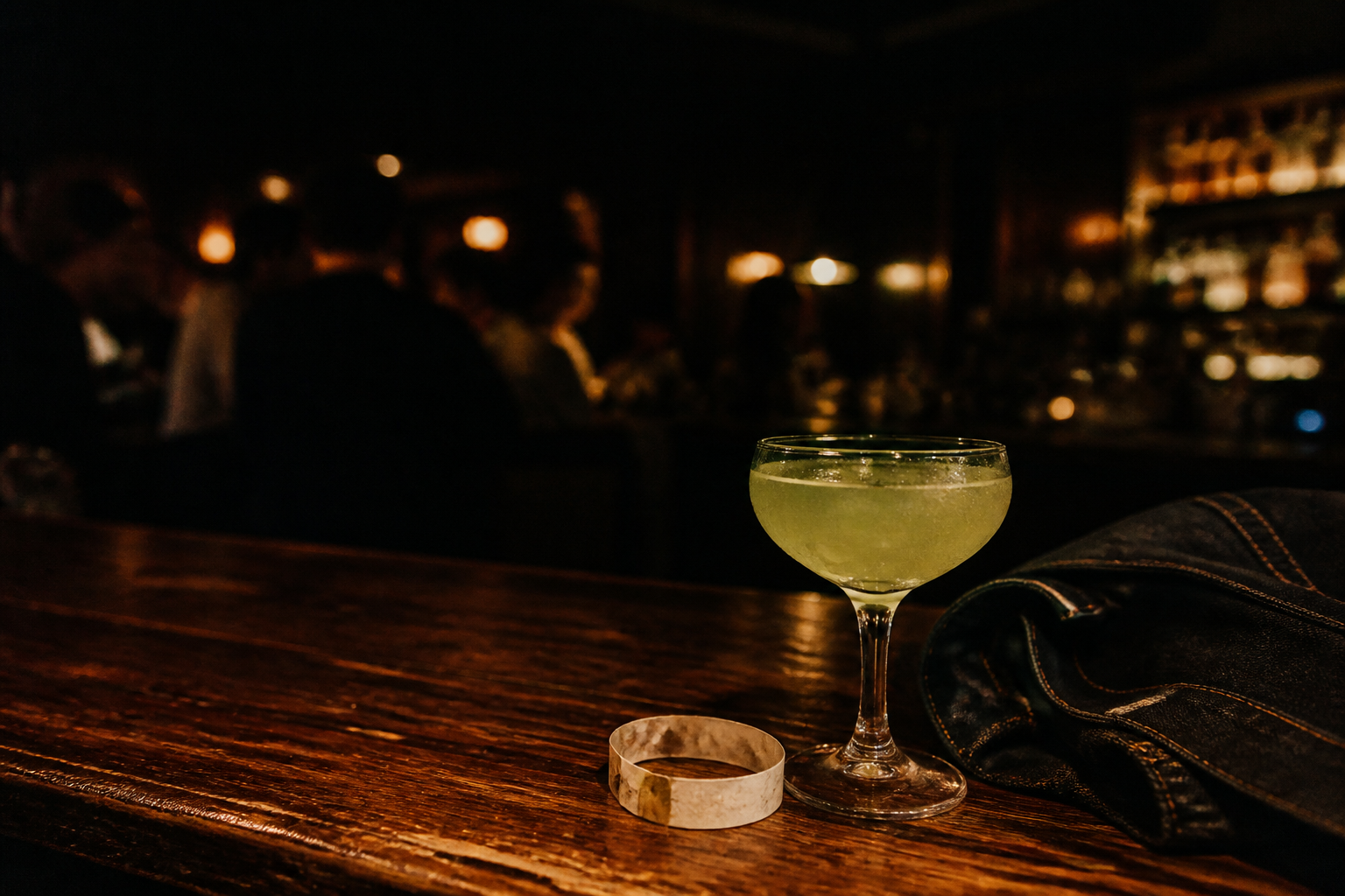 A pale green cocktail on a worn wooden bar beside a venue wristband and dark denim jacket.
